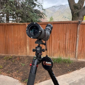 Op de afbeelding: Een zwarte spotting scope gemonteerd op een zwart statief met rode accenten. De scope is gericht op een bergketen op de achtergrond. Het statief heeft een gewatteerde handgreep en een "Glass Guardian" logo.