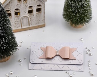 May include: A white envelope with a pink bow and pearl embellishment. The envelope is decorated with a white embossed pattern. The envelope is surrounded by white pearls.
