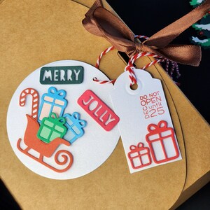 May include: A brown cardboard box with a white circle attached to the front. The circle has a red sleigh with candy canes and presents, and the words "Merry" and "Jolly". A white tag with red text says "Do Not Open Until Dec 25" and has two red presents. The box has a brown ribbon tied around it.