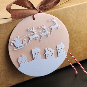 May include: A round, light pink Christmas ornament with a white silhouette of Santa Claus in a sleigh pulled by reindeer over a cityscape. The ornament is tied with a brown ribbon and red and white twine.