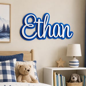 May include: A wall-mounted wooden sign featuring the name Ethan in a cursive script font. The sign consists of a white top layer with a blue backing layer, creating a layered effect. The letters are connected and display a smooth, clean finish.
