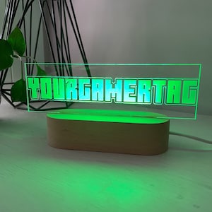 May include: A green illuminated gamer tag sign with the text "YOURGAMERTAG" in a pixelated font. The sign is made of clear acrylic and sits on a wooden base. The light emits a green glow.
