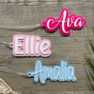 May include: Three personalized name tags with white lettering on a wood background. The names are Ava in pink, Ellie in light pink, and Amalia in light blue.