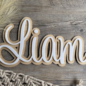 May include: A custom name sign featuring the name LIAM in white, raised, cursive lettering layered over a light-toned wood backing. The sign has a smooth, rounded contour that follows the shape of the letters, creating a layered effect.