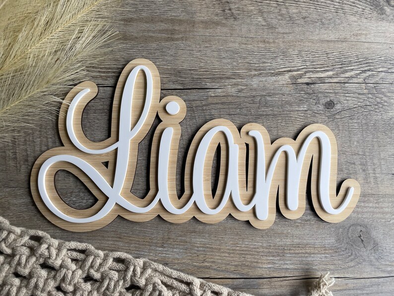 May include: A custom name sign featuring the name LIAM in white, raised, cursive lettering layered over a light-toned wood backing. The sign has a smooth, rounded contour that follows the shape of the letters, creating a layered effect.