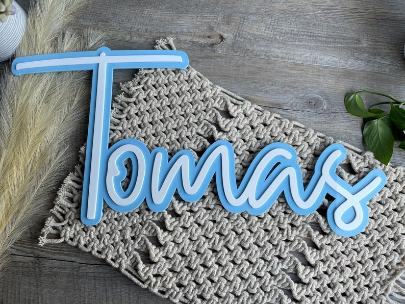 May include: A light blue wooden name sign with white lettering spelling out the name "Tomas". The sign is on a beige crocheted surface.