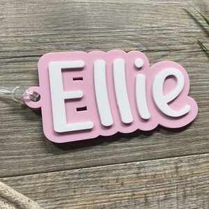 May include: Pink personalized name tag with the name "Ellie" in white lettering. The tag has a clear plastic loop for attaching to a bag or other item.