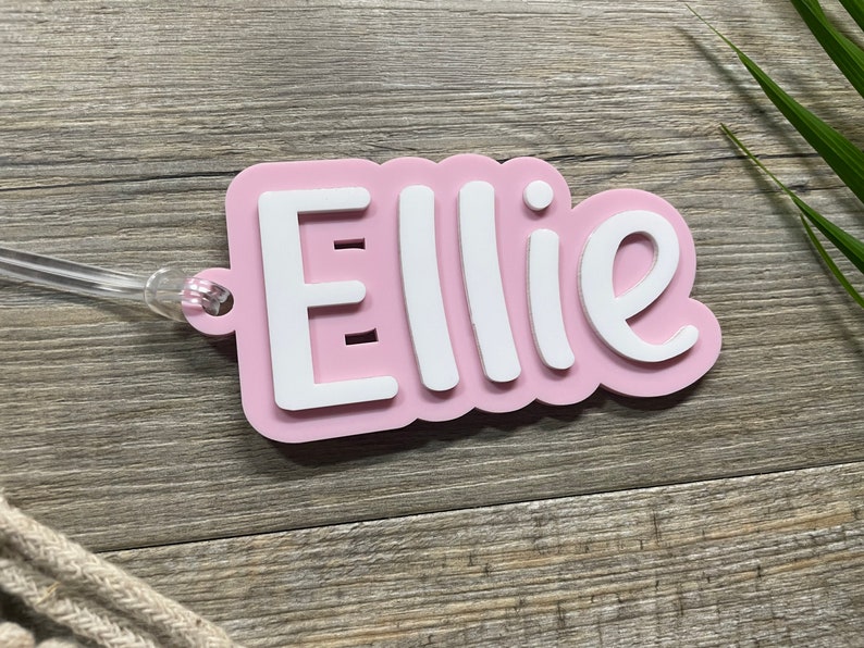 May include: Pink personalized name tag with the name "Ellie" in white lettering. The tag has a clear plastic loop for attaching to a bag or other item.