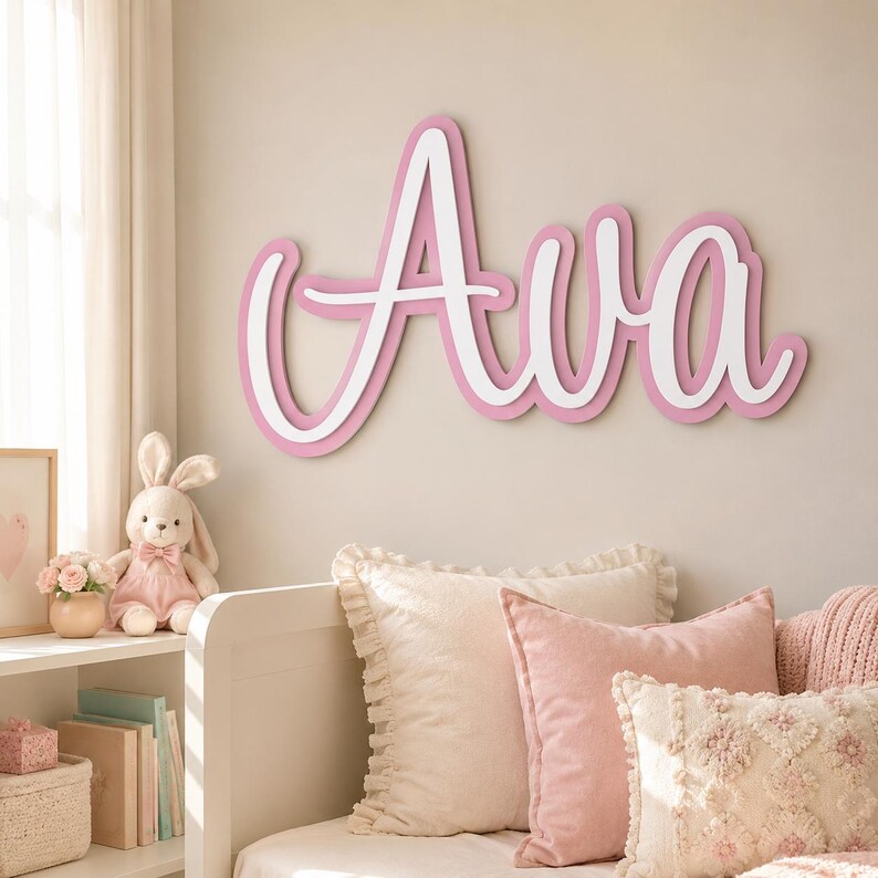 May include: A wall-mounted sign featuring the name Ava in a cursive script font. The lettering consists of a white top layer with a pink border, creating a layered effect. The sign is mounted on a plain wall above a bed with pillows.