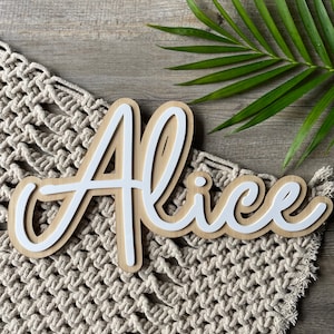 May include: White wooden name sign with the name "Alice" written in a cursive script. The sign is on a beige crocheted surface.