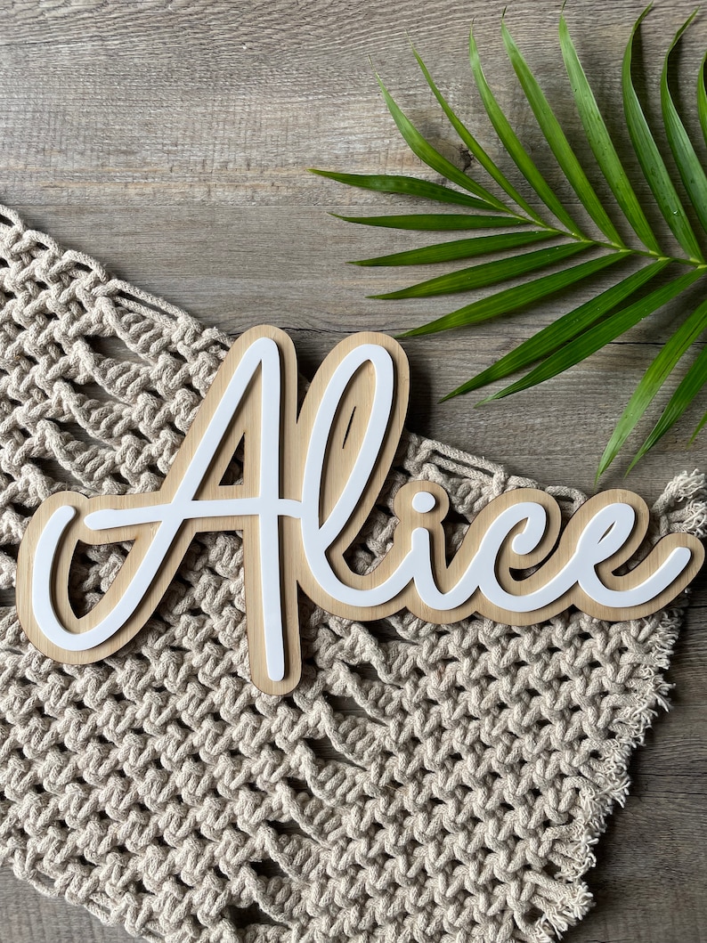 May include: White wooden name sign with the name "Alice" written in a cursive script. The sign is on a beige crocheted surface.