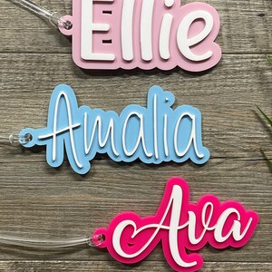 May include: Three personalized name tags with white lettering. The top tag is pink with the name "Ellie", the middle tag is light blue with the name "Amalia", and the bottom tag is hot pink with the name "Ava".