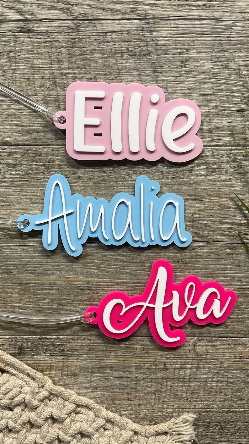 May include: Three personalized name tags with white lettering. The top tag is pink with the name "Ellie", the middle tag is light blue with the name "Amalia", and the bottom tag is hot pink with the name "Ava".