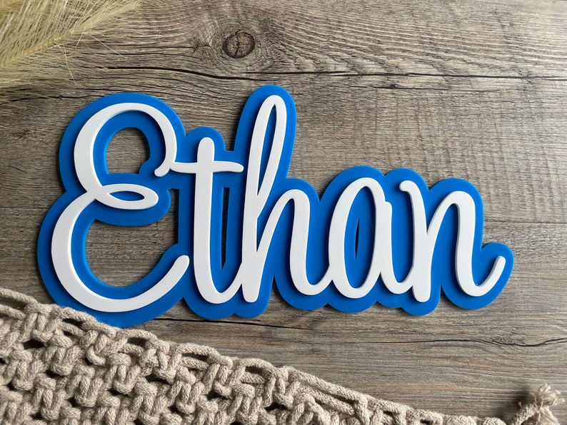 May include: A custom name sign featuring the name Ethan in white script lettering layered over a solid blue background. The sign has a smooth, clean finish with a distinct contrast between the white letters and the blue backing plate.