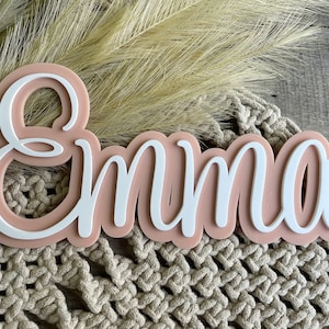 May include: A custom name sign featuring the name Emma in white script lettering layered over a solid, rounded, light peach-colored acrylic backing. The sign has a smooth, matte finish and a clean, modern aesthetic with a raised 3D effect.