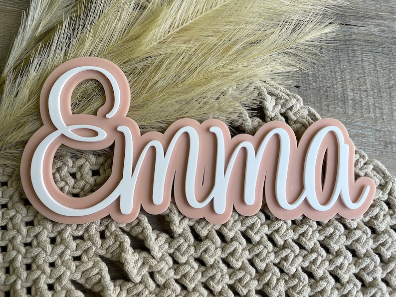 May include: A custom name sign featuring the name Emma in white script lettering layered over a solid, rounded, light peach-colored acrylic backing. The sign has a smooth, matte finish and a clean, modern aesthetic with a raised 3D effect.