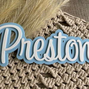 May include: A custom name sign featuring the name Preston in white, raised, cursive lettering layered over a light blue, cloud-shaped backing. The sign has a smooth, matte finish and a layered 3D design that creates depth and visual contrast.