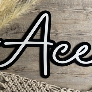 May include: Black and white cursive script name sign with the name "Ace" written in white letters with a black outline. The sign is on a wood background.