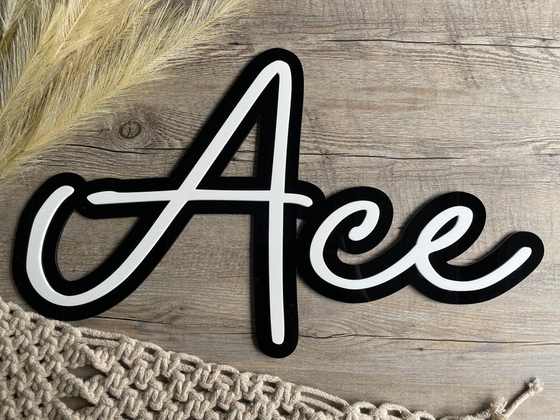 May include: Black and white cursive script name sign with the name "Ace" written in white letters with a black outline. The sign is on a wood background.