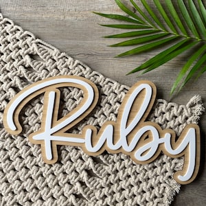 May include: White wooden name sign with the name "Ruby" written in a cursive font. The sign is on a beige crocheted surface.