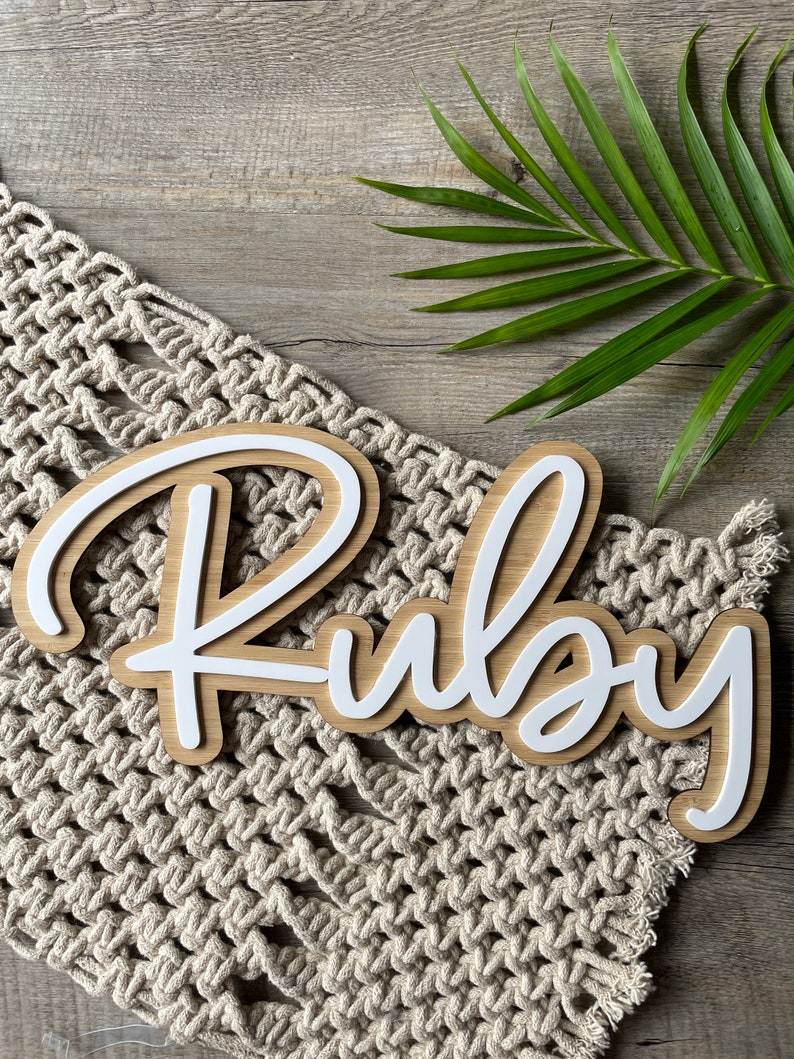 May include: White wooden name sign with the name "Ruby" written in a cursive font. The sign is on a beige crocheted surface.