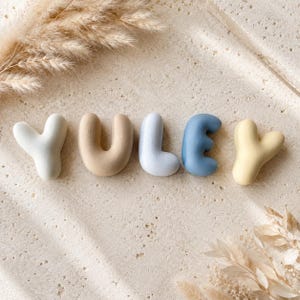 May include: Five individual three-dimensional letters spelling the word YULEY. The letters are smooth, rounded, and matte in finish, featuring a colour palette of off-white, beige, light blue, medium blue, and pale yellow, arranged in a horizontal row.