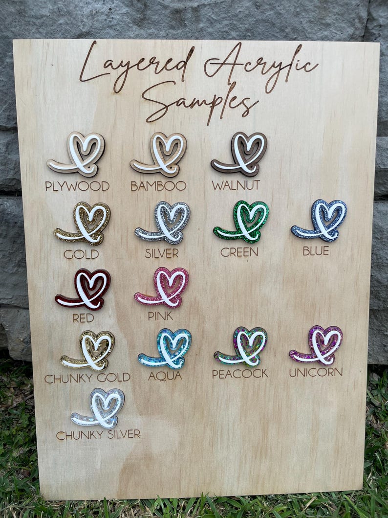 May include: A light wood display board titled Layered Acrylic Samples featuring fourteen heart-shaped icons in various finishes including plywood, bamboo, walnut, gold, silver, green, blue, red, pink, chunky gold, aqua, peacock, unicorn, and chunky silver.