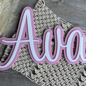 May include: A custom name sign featuring the name Ava in a white script font layered over a larger, matching dusty pink backing. The letters are connected in a continuous cursive style, creating a dimensional effect with the two-tone color scheme.
