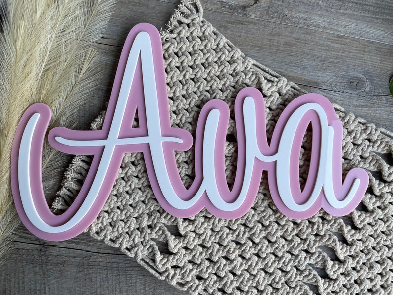 May include: A custom name sign featuring the name Ava in a white script font layered over a larger, matching dusty pink backing. The letters are connected in a continuous cursive style, creating a dimensional effect with the two-tone color scheme.