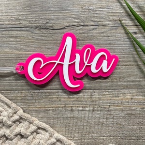 May include: Pink acrylic luggage tag with the name "Ava" written in white cursive lettering.