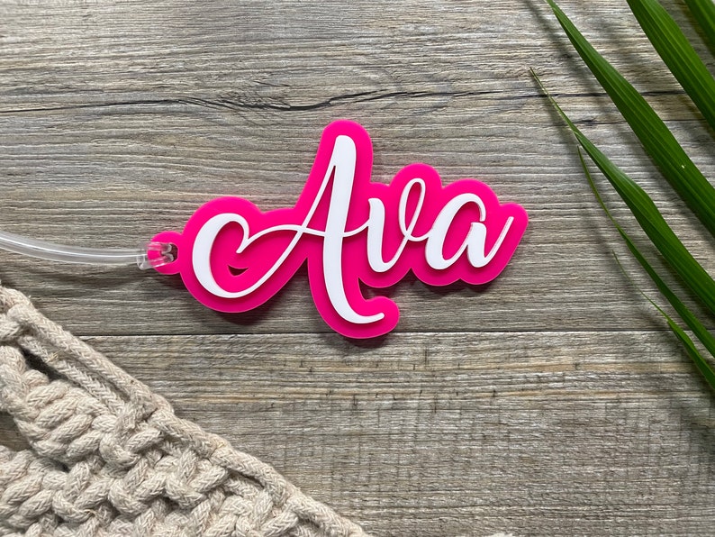 May include: Pink acrylic luggage tag with the name "Ava" written in white cursive lettering.