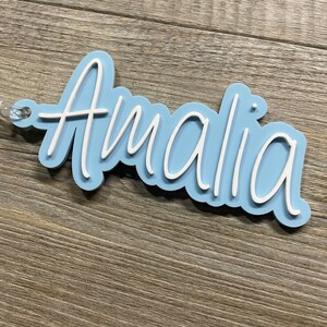 May include: A light blue acrylic name tag with the name "Amalia" written in white cursive lettering.