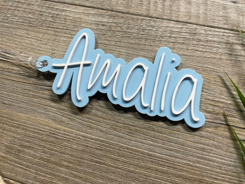 May include: A light blue acrylic name tag with the name "Amalia" written in white cursive lettering.