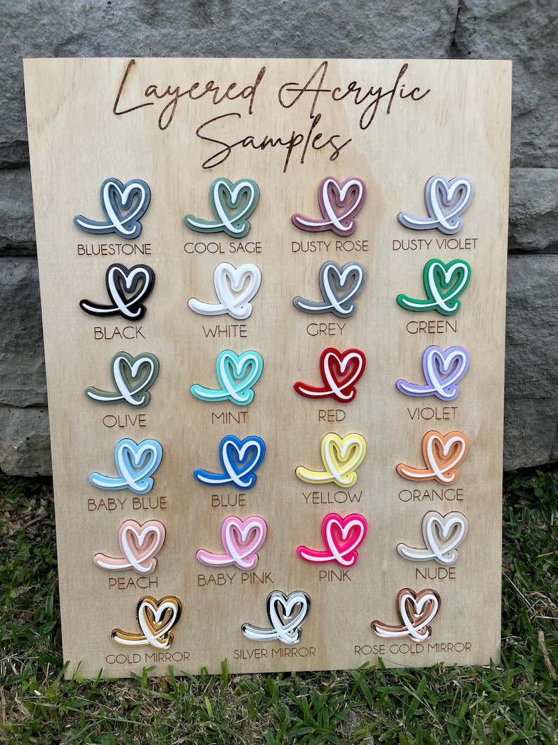 May include: A wooden display board titled Layered Acrylic Samples featuring twenty-three heart-shaped acrylic pieces in various colors including bluestone, cool sage, dusty rose, black, white, grey, green, olive, mint, red, violet, baby blue, blue, yellow, orange, peach, baby pink, pink, nude, gold mirror, silver mirror, and rose gold mirror.