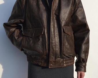 May include: A brown leather bomber jacket with two front pockets and a ribbed hem. The jacket has a collared neckline and a zipper closure. The leather has a distressed appearance. The jacket is worn over a dark gray turtleneck.
