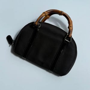 May include: Black handbag with a bamboo handle. The bag has a rounded shape with a zipper closure and a small leather pull tab. The handle is made of curved bamboo, and the bag has black leather accents.