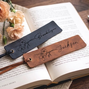 May include: Two rectangular leather bookmarks, one black and one tan, each featuring a laser-engraved floral illustration and a personalized name in cursive script. The black bookmark reads Brooklyn, and the tan bookmark reads Jackson.