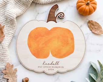 May include: A light-colored wooden pumpkin-shaped plaque featuring a central orange painted pumpkin design. The plaque includes the printed text Kendall, our little bumpkin, and 2024, with a dark brown wooden stem attached to the top edge.