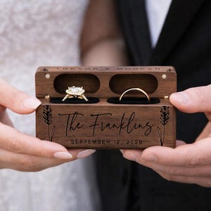 May include: A wooden ring box with two compartments, one holding a diamond engagement ring and the other a gold wedding band. The box is engraved with "The Franklings" and the date "September 12, 2020".