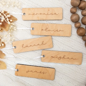 Puede incluir: Cinco etiquetas de regalo de madera con nombres escritos en cursiva. Los nombres son Veronica, Oliver, Hannah, Meghan y James. Las etiquetas están sobre una superficie de punto blanco con una guirnalda de cuentas marrones.