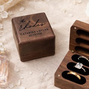 May include: Two wooden ring boxes, one closed and one open, displaying rings. The closed box is engraved with "The Rhodes", names, and a date. The open box holds three rings: a gold band and two diamond rings. The background includes flowers.
