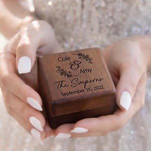 May include: A small, square, wooden ring box held in hands. The box is engraved with the names "Cole & Amy", the family name "The Simpsons", and the date "September 15, 2022". Floral designs are also engraved on the box.