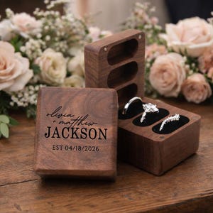 以下が含まれることがあります： 濃い木製のリングボックス。一つは開いており、黒いベルベットの上に婚約指輪が飾られています。もう一つは閉じており、「Olivia + Matthew JACKSON」の名前と「EST 04/18/2026」の日付が刻まれています。