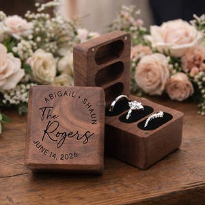May include: Wooden ring boxes with a wedding theme. One box is engraved with "Abigail + Shaun The Rogers JUNE 14, 2026". The other box holds two rings. The boxes are on a wooden surface with flowers in the background.