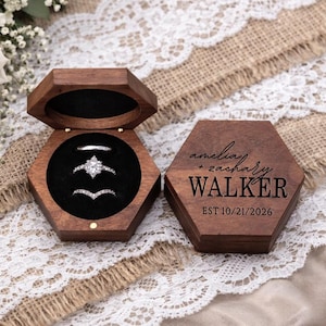 May include: A wooden hexagonal ring box, open to reveal three rings with sparkling stones, sits on a lace-trimmed surface. The box is next to a closed box with the names "amelia + zachary WALKER" and the date "EST 10/21/2026" engraved on it.