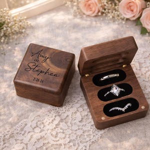 May include: A wooden ring box with the names "Amy and Stephen" and the date "2.26.26" engraved on the lid. The open box displays three rings in black velvet slots. The box is made of dark wood.