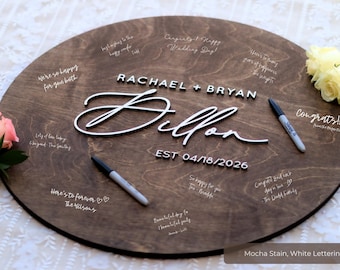 May include: A round wooden wedding sign with the names "Rachael + Bryan" and "Dillon" in white lettering. The date "EST 04/18/2026" is also displayed. The sign is stained mocha and has handwritten messages.