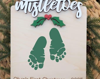 Puede incluir: Un adorno navideño de madera con la palabra "mistletoes" en escritura blanca. Presenta huellas verdes, hojas de acebo y el texto "Olivia's First Christmas - 2025". El adorno está sobre un fondo tejido.