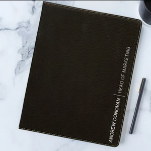 May include: A black leather-bound notebook with a gold stitched border. The notebook has the text "ANDREW DONOVAN | HEAD OF MARKETING" embossed in white on the front cover.