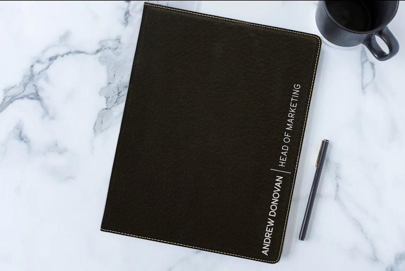 May include: A black leather-bound notebook with a gold stitched border. The notebook has the text "ANDREW DONOVAN | HEAD OF MARKETING" embossed in white on the front cover.